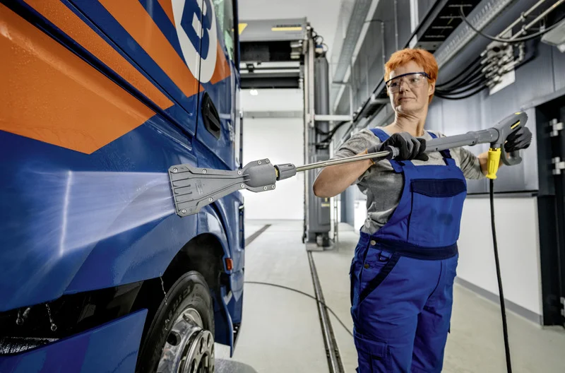Person reinigt Bus mit Kärcher Hochdruckreiniger in Werkstatt. Bus hat blaue und orangefarbene Lackierung.