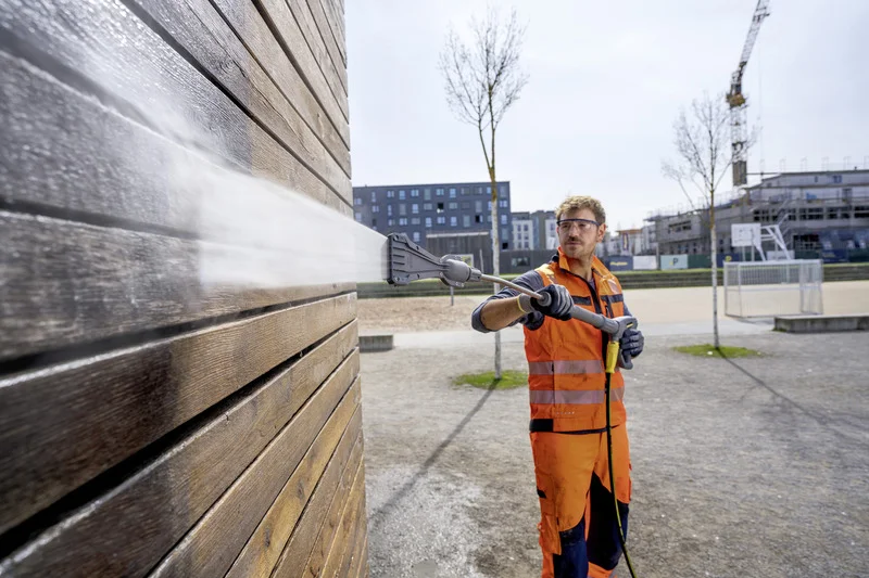 Arbeiter in orangefarbener Schutzkleidung reinigt Holzfassade mit Kärcher Hochdruckreiniger im Freien, Baustelle im Hintergrund.